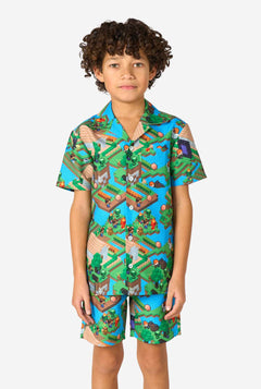 A young boy wearing a colorful summer outfit featuring a Minecraft Overworld print, showcasing pixelated landscapes, characters, and elements from the iconic game. The short-sleeve shirt and matching shorts highlight a vibrant design inspired by the Minecraft universe.