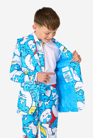 Teen boy wearing a vibrant Smurfs kids suit with colorful cartoon character prints in blue, red, yellow, and white, paired with a matching tie and white shirt, smiling against a plain background. Holding one side of the jacket open.