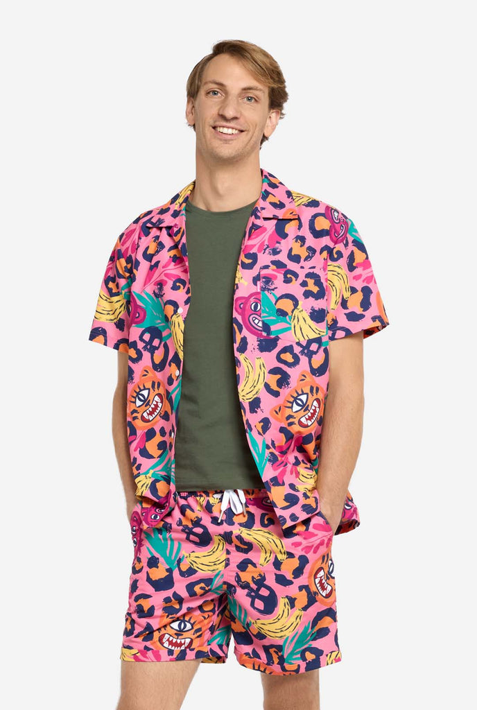 A man wearing a vibrant two-piece summer outfit featuring a bold tropical print. The short-sleeve button-up shirt and matching shorts showcase a lively design with neon pink, orange, and yellow leopard prints, bananas, green palm leaves, and quirky cartoon faces. The outfit is styled casually with a green T-shirt underneath and is perfect for summer parties, vacations, or poolside fun. 