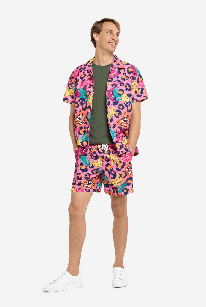 A man wearing a vibrant two-piece summer outfit featuring a bold tropical print. The short-sleeve button-up shirt and matching shorts showcase a lively design with neon pink, orange, and yellow leopard prints, bananas, green palm leaves, and quirky cartoon faces. The outfit is styled casually with a green T-shirt underneath and is perfect for summer parties, vacations, or poolside fun. Man is looking sideways.