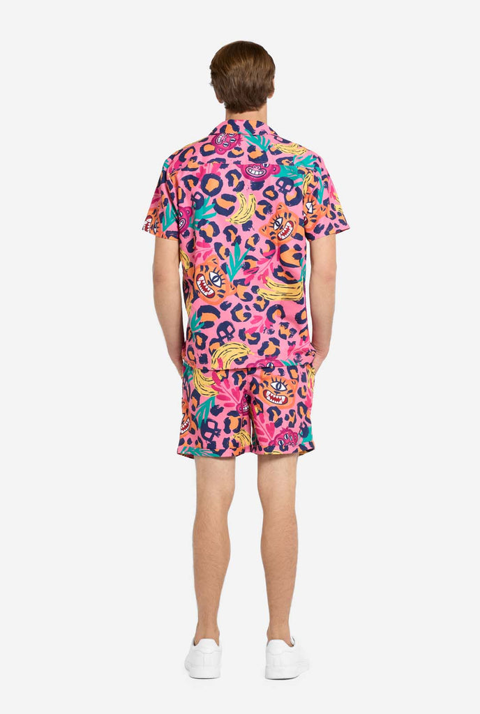 A man wearing a vibrant two-piece summer outfit featuring a bold tropical print. The short-sleeve button-up shirt and matching shorts showcase a lively design with neon pink, orange, and yellow leopard prints, bananas, green palm leaves, and quirky cartoon faces. The outfit is styled casually with a green T-shirt underneath and is perfect for summer parties, vacations, or poolside fun. View from the back.
