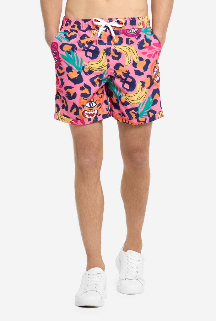 A man wearing a vibrant two-piece summer outfit featuring a bold tropical print. The short-sleeve button-up shirt and matching shorts showcase a lively design with neon pink, orange, and yellow leopard prints, bananas, green palm leaves, and quirky cartoon faces. The outfit is styled casually with a green T-shirt underneath and is perfect for summer parties, vacations, or poolside fun. Close up of the shorts.
