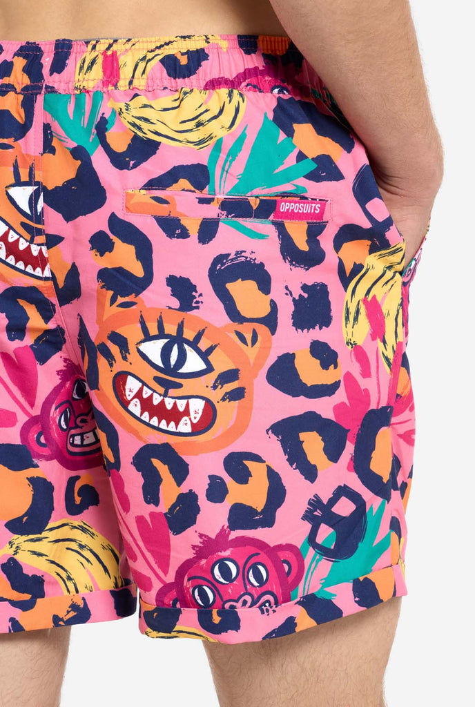 A man wearing a vibrant two-piece summer outfit featuring a bold tropical print. The short-sleeve button-up shirt and matching shorts showcase a lively design with neon pink, orange, and yellow leopard prints, bananas, green palm leaves, and quirky cartoon faces. The outfit is styled casually with a green T-shirt underneath and is perfect for summer parties, vacations, or poolside fun. Back of short close up.