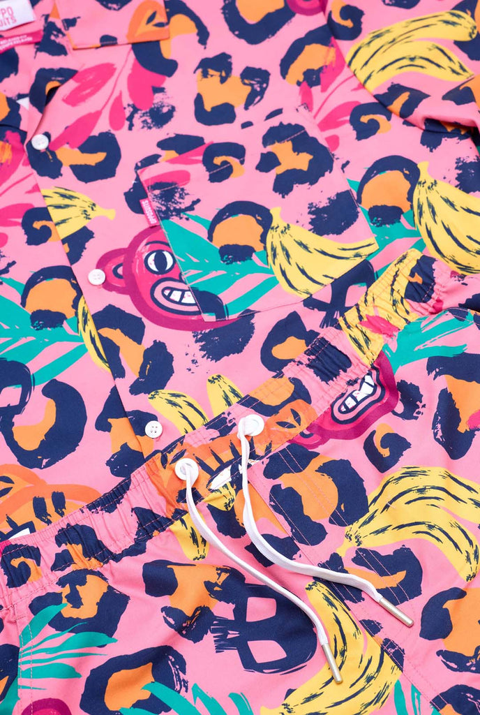 Image of a vibrant two-piece summer outfit featuring a bold tropical print. The short-sleeve button-up shirt and matching shorts showcase a lively design with neon pink, orange, and yellow leopard prints, bananas, green palm leaves, and quirky cartoon faces. The outfit is styled casually with a green T-shirt underneath and is perfect for summer parties, vacations, or poolside fun. Close-up.