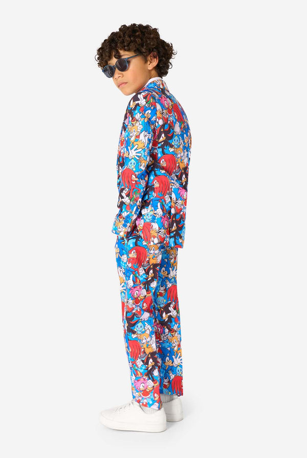 Young boy wearing sunglasses and a vibrant Sonic teen suit featuring colorful prints of Sonic the Hedgehog characters including Sonic, Knuckles, Tails, and Amy, paired with a matching tie and white shirt. Side view.