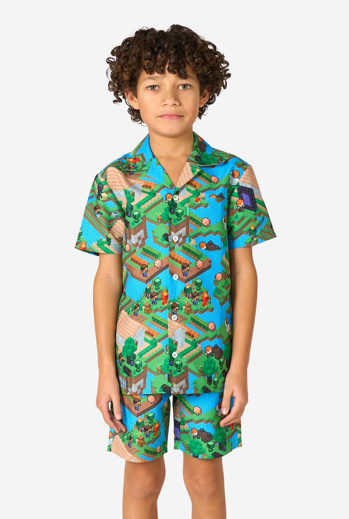 A young boy wearing a colorful summer outfit featuring a Minecraft Overworld print, showcasing pixelated landscapes, characters, and elements from the iconic game. The short-sleeve shirt and matching shorts highlight a vibrant design inspired by the Minecraft universe. 