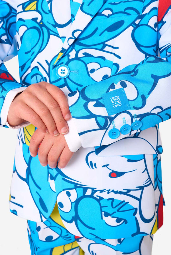 Young boy wearing a colorful Smurfs-themed kids suit featuring vibrant blue cartoon character prints, including jacket, pants, and matching tie, smiling against a plain white background. Sleeves close up.