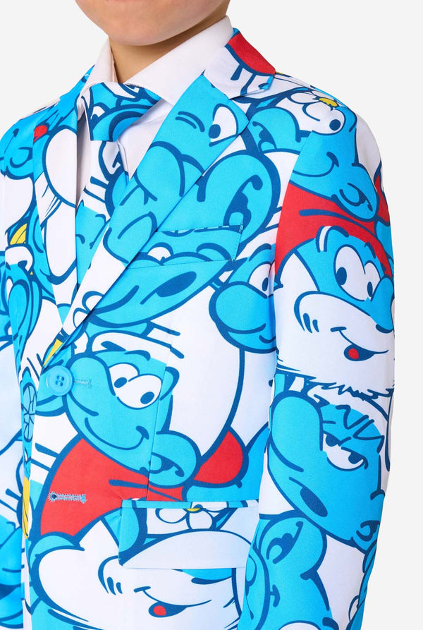 Young boy wearing a colorful Smurfs-themed kids suit featuring vibrant blue cartoon character prints, including jacket, pants, and matching tie, smiling against a plain white background. Chest close up.