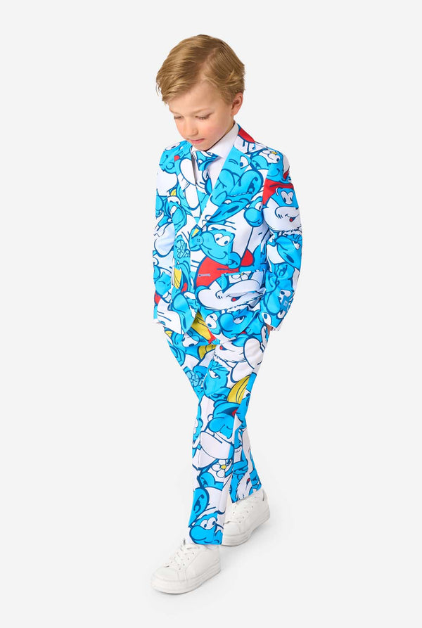 Young boy wearing a colorful Smurfs-themed kids suit featuring vibrant blue cartoon character prints, including jacket, pants, and matching tie, smiling against a plain white background. Looking to the ground.