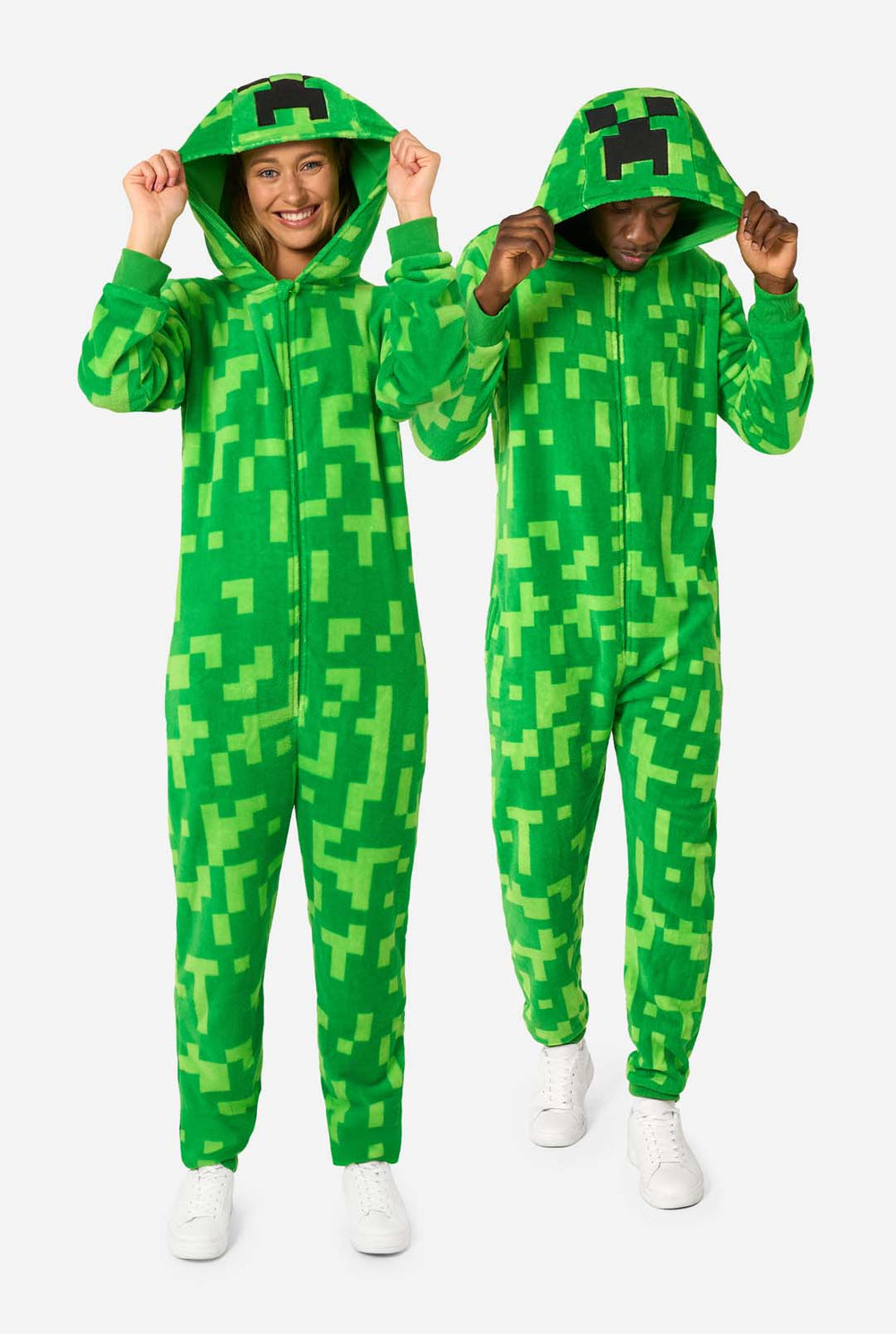 Two people wearing green pixelated onesies designed to look like the Minecraft Creeper character. The onesies have hoods with the Creeper’s iconic black pixel face. The man is holding his hood and looking down, while the women is smiling at the camera with one hand on their hip