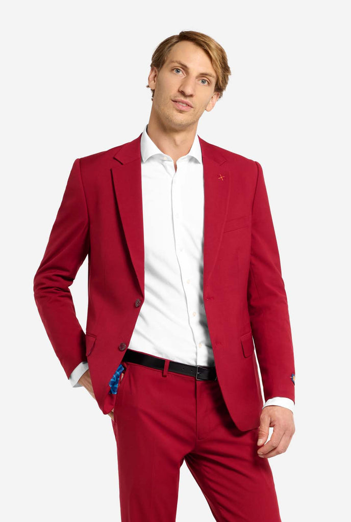 Image with light grey background and man wearing Bright Burgundy men’s suit and white shirt. Suits consists of blazer and pants. Man looking in the camera