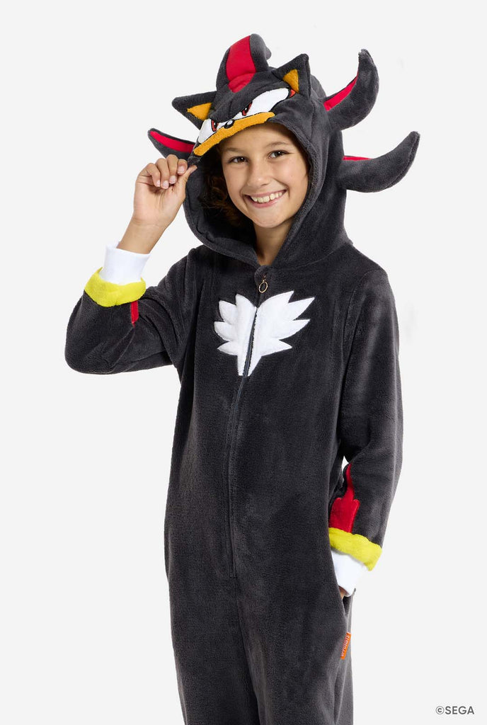 A kid wearing a plush black kids onesie inspired by the popular hedgehog character Shadow, featuring spiky red and black accents on the hood, a white chest emblem, and yellow wrist details. The onesie is made from soft fleece material, offering comfort and a playful design for cosplay, lounging, or themed events. The person smiles while the hood is on his head. Perfect for fans of gaming or animated characters.