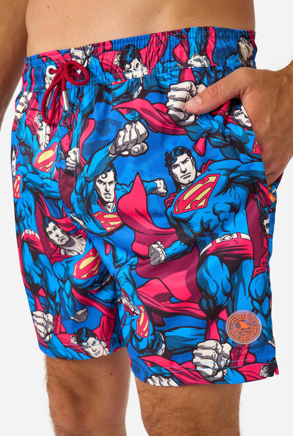 Man wearing a Men's Superman-themed swim shorts featuring a bold comic book design with multiple action poses of the Man of Steel. Perfect for superhero fans looking to make a splash.  Close-up front.