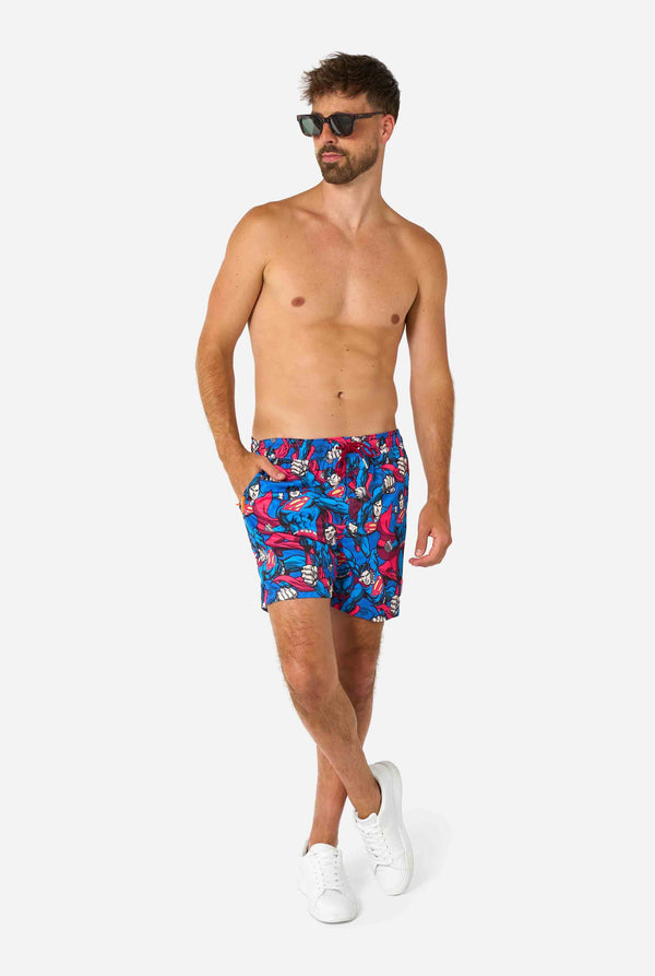 Man wearing a Men's Superman-themed swim shorts featuring a bold comic book design with multiple action poses of the Man of Steel. Perfect for superhero fans looking to make a splash. Full body image and man wearing sunglasses.