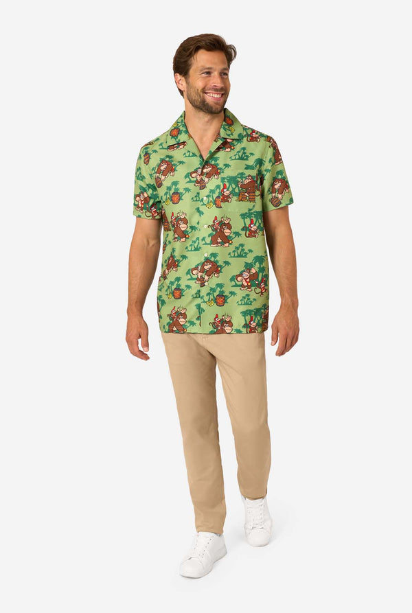 Image of a man wearing a Donkey Kong themed Hawaiian shirt. The shirt has a light green background and contains dark green palm trees, and multiple images of the character Donkey Kong, from Super Mario. The man is looking to the side, full body image.