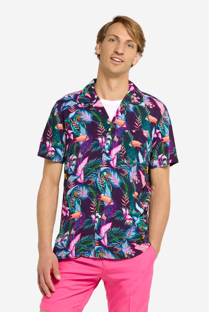 A man is wearing a purple Hawaiian shirt with a tropical print, resembling the jungle night live. Print consists of parrots, snakes, leaves and flowers. Also wearing pink shorts. He is looking in the camera and holding holding one hand in the pocket.