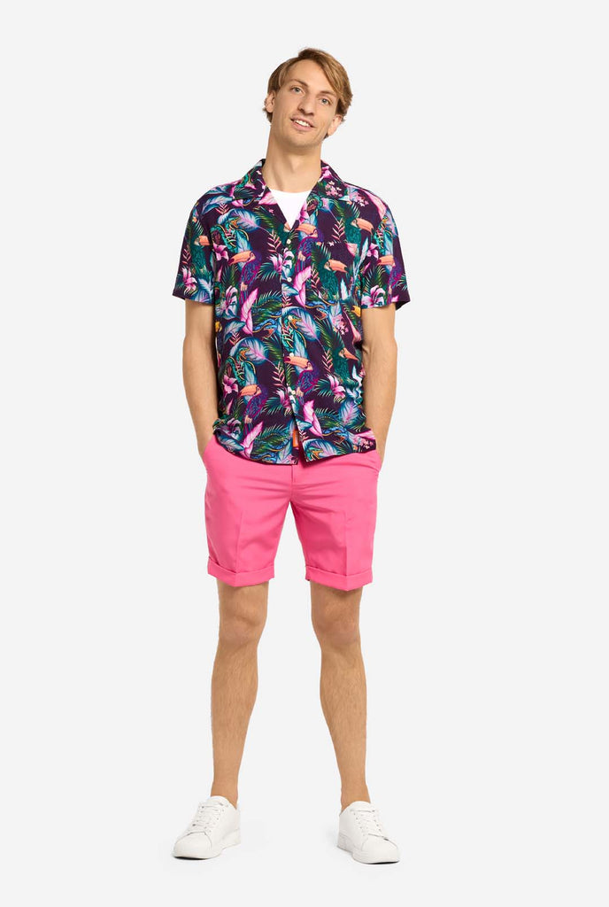 A man is wearing a purple Hawaiian shirt with a tropical print, resembling the jungle night live. Print consists of parrots, snakes, leaves and flowers. Also wearing pink shorts. He is looking in the camera and holding his hands in the pockets.