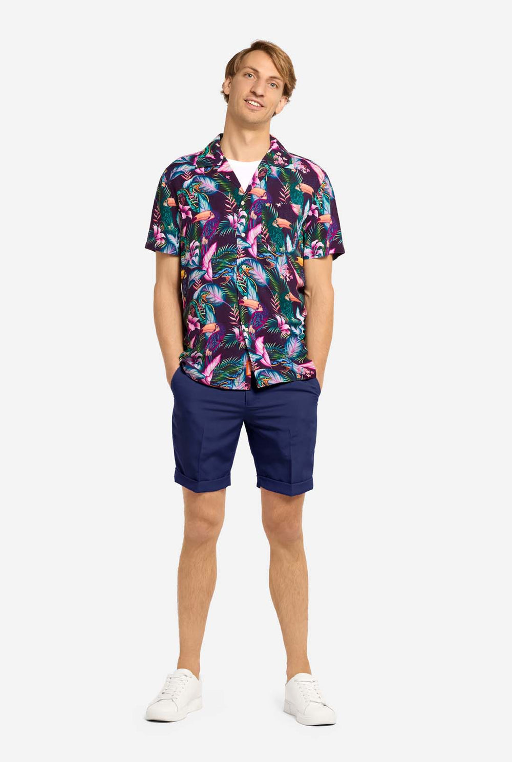 A man wearing a colorful short-sleeve Hawaiian shirt with a vibrant jungle pattern featuring tropical birds, leaves, and flowers in shades of pink, blue, green, and purple. He has light brown hair, is smiling slightly, and is standing with one hand in his pocket against a plain light background.