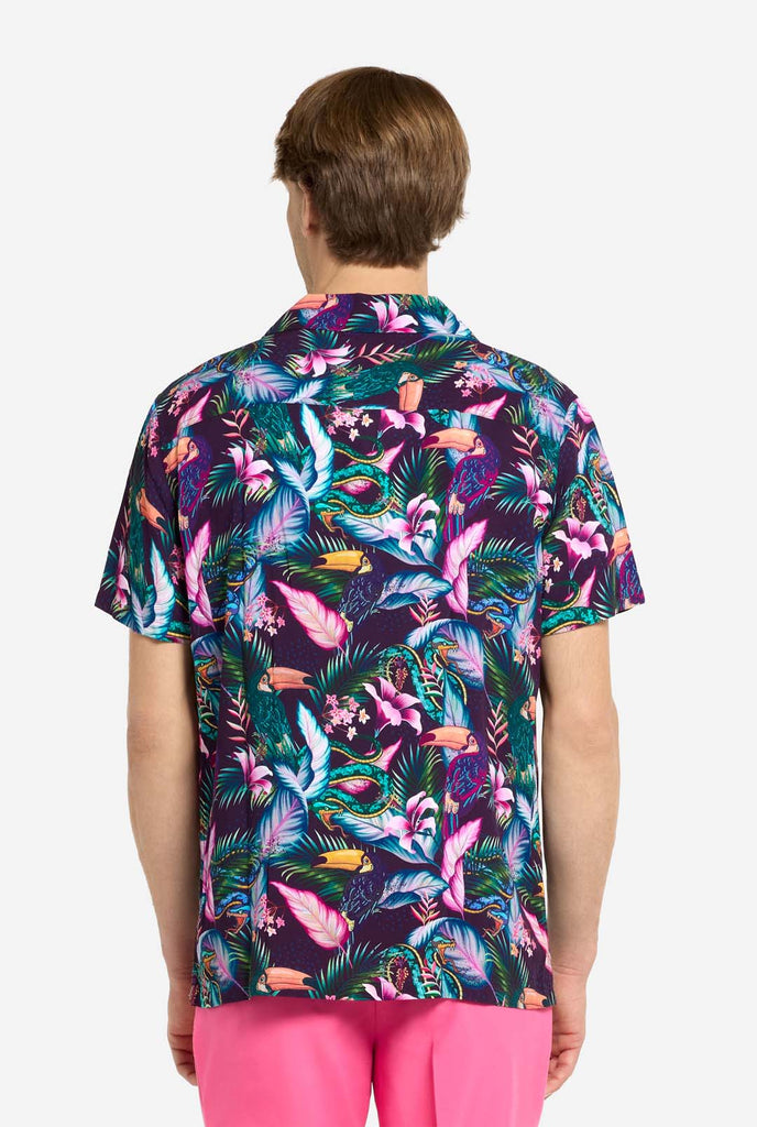 A man is wearing a purple Hawaiian shirt with a tropical print, resembling the jungle night live. Print consists of parrots, snakes, leaves and flowers. Also wearing pink shorts. View from the back.