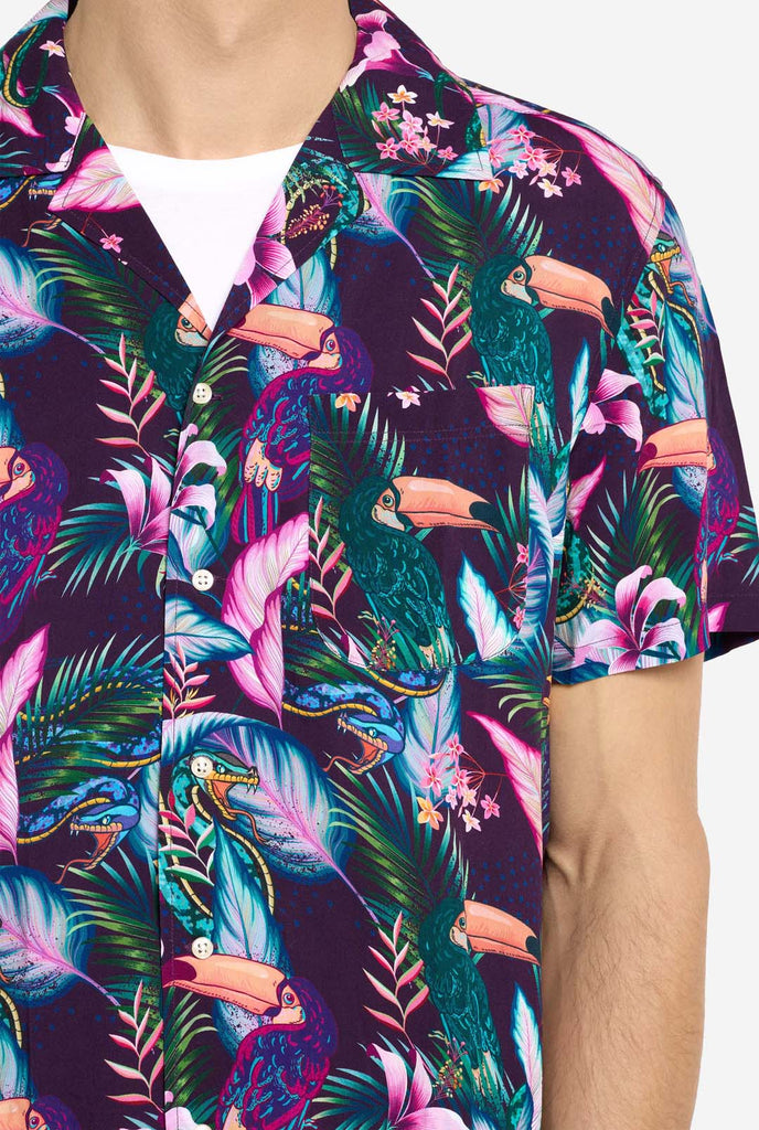 A man is wearing a purple Hawaiian shirt with a tropical print, resembling the jungle night live. Print consists of parrots, snakes, leaves and flowers. Chest close-up.