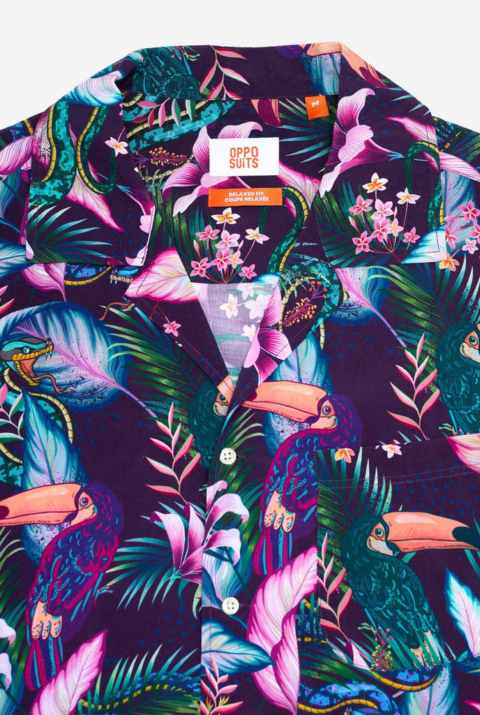 A man is wearing a purple Hawaiian shirt with a tropical print, resembling the jungle night live. Print consists of parrots, snakes, leaves and flowers. Chest close-up.