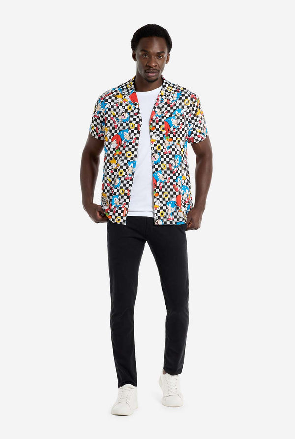 A man is wearing a black and white checkered Hawaiian shirt with characters from the Sonic the Hedgehog franchise and golden rings on it. Also wearing black pants. He is looking in the camera and holding the bottom of the shirt.