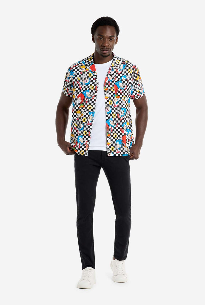 A man is wearing a black and white checkered Hawaiian shirt with characters from the Sonic the Hedgehog franchise and golden rings on it. Also wearing black pants. He is looking in the camera and holding the bottom of the shirt.