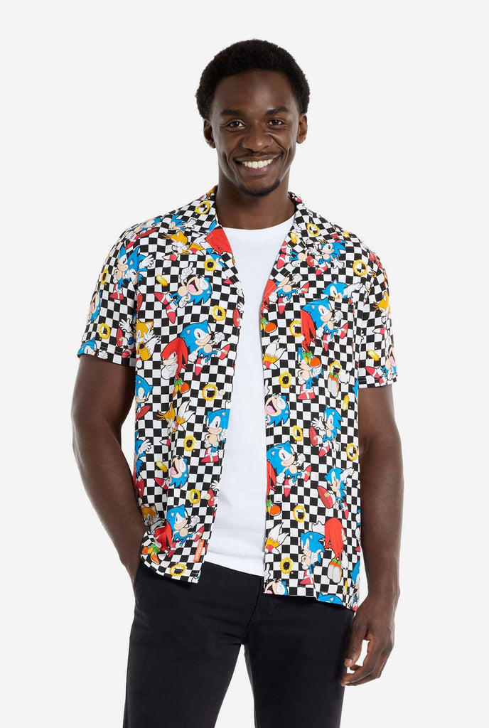 A man is wearing a black and white checkered Hawaiian shirt with characters from the Sonic the Hedgehog franchise and golden rings on it. Also wearing black pants. He is smiling in the camera and holding the bottom of the shirt.