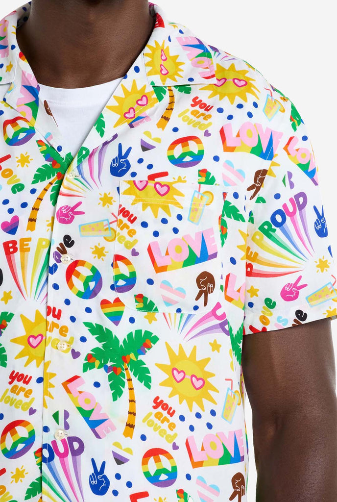 A man is wearing a white Hawaiian shirt with a rainbow pride print. The Print consists of peace symbols, hearts, and other icons. Chest close-up.