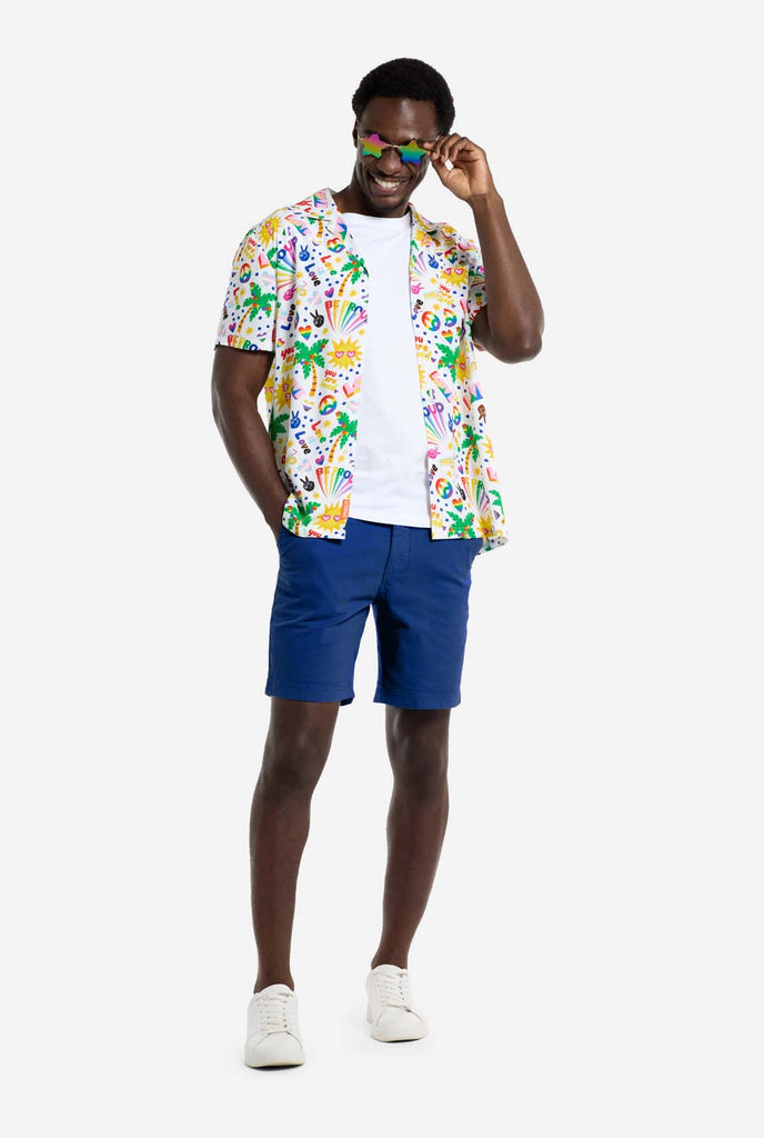 A man is wearing a white Hawaiian shirt with a rainbow pride print. The Print consists of peace symbols, hearts, and other icons. He is also wearing blue pants. He is smiling and wearing star-shaped sunglasses.