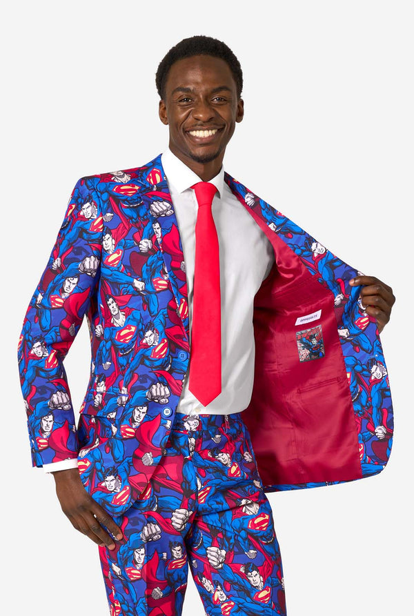 A man wearing a Superman themed suit with a red tie and a white shirt on a white background. Holding his jacket open