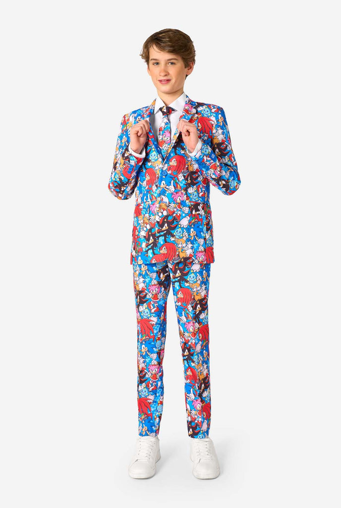 Teen boy wearing a vibrant Sonic teen suit featuring colorful prints of Sonic the Hedgehog characters including Sonic, Knuckles, Tails, and Amy, paired with a matching tie and white shirt. Full body image.