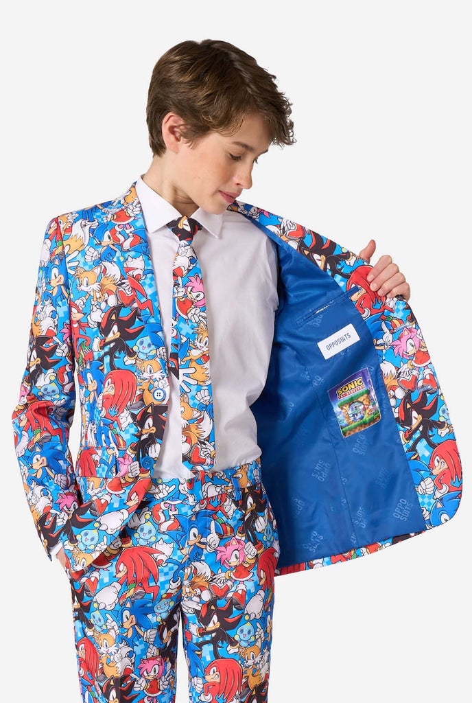 Teen boy wearing a vibrant Sonic teen suit featuring colorful prints of Sonic the Hedgehog characters including Sonic, Knuckles, Tails, and Amy, paired with a matching tie and white shirt. Holding one side of the jacket open.