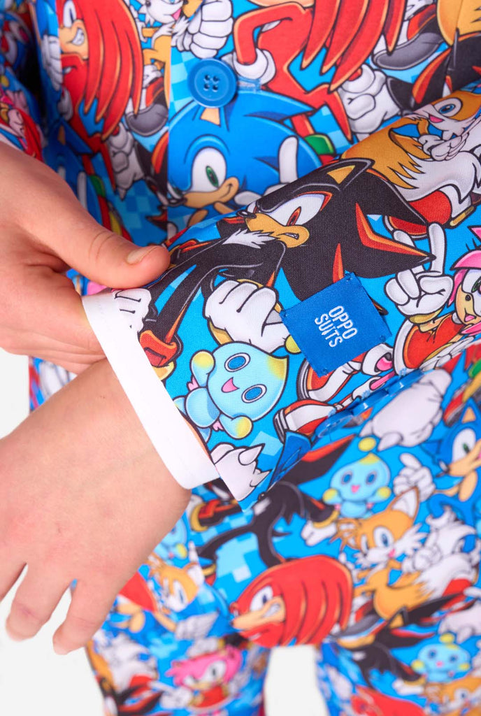 Teen boy wearing a vibrant Sonic teen suit featuring colorful prints of Sonic the Hedgehog characters including Sonic, Knuckles, Tails, and Amy, paired with a matching tie and white shirt. Sleeve close up.
