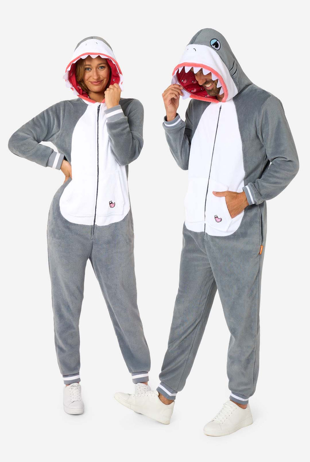 A man and women are wearing a grey and white shark adult onesie featuring playful hood details and cozy, soft material, perfect for lounging or costume events. Both are wearing their hoods on their head.