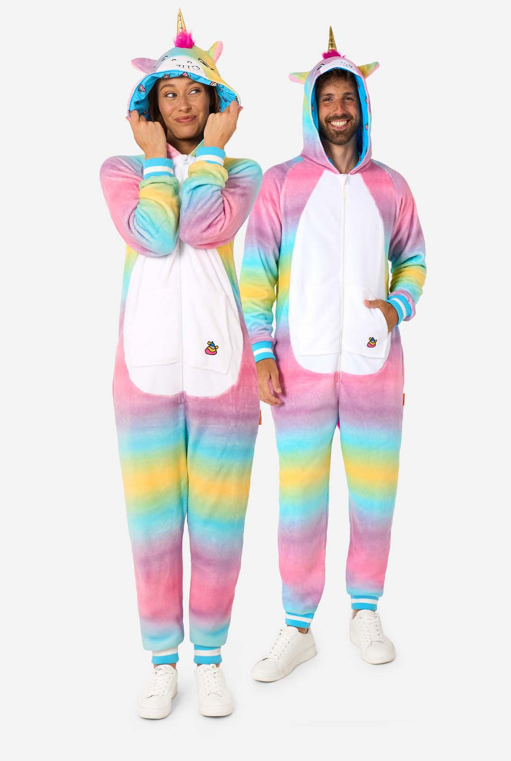 A man and women are wearing a colorful adult onesie designed as a rainbow unicorn. The onesie features a vibrant gradient of pastel colors, a golden horn, and playful unicorn details, perfect for cozy lounging or costume parties. The men and women both have their hood on.