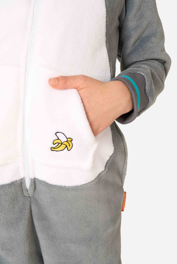 Child wearing a cozy unisex kids onesie designed as a lemur, featuring grey and white plush fabric, a hood with a cartoon lemur face and ears, and a playful banana print on the pocket. Holding a hand in a pocket.