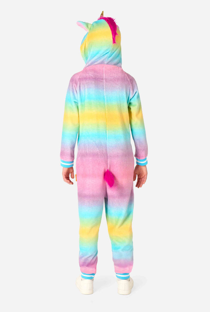 A smiling kid is wearing a white, pink, blue and yellow kids onesie designed as a unicorn. The Unicorn onesie features a hood resembling a unicorn head and other playfull details. View from the back, full body.