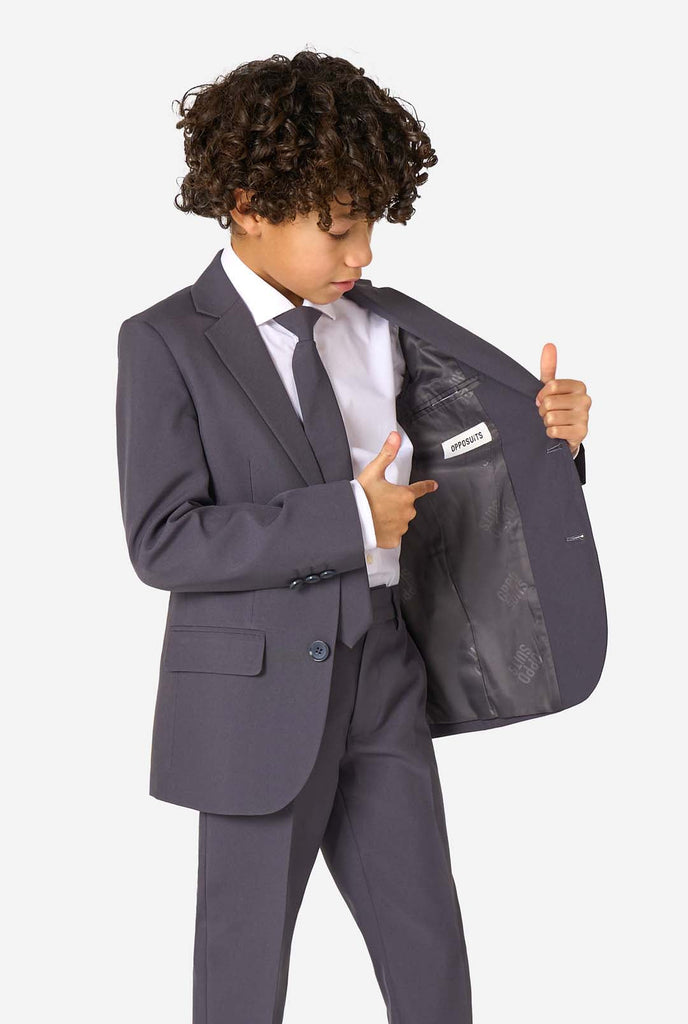 Kid wearing a stylish charcoal colored grey suit with a white dress shirt, smiling confidently. Perfect formal outfit for boys' special occasions, weddings, and parties. Holding one side of the jacket open.