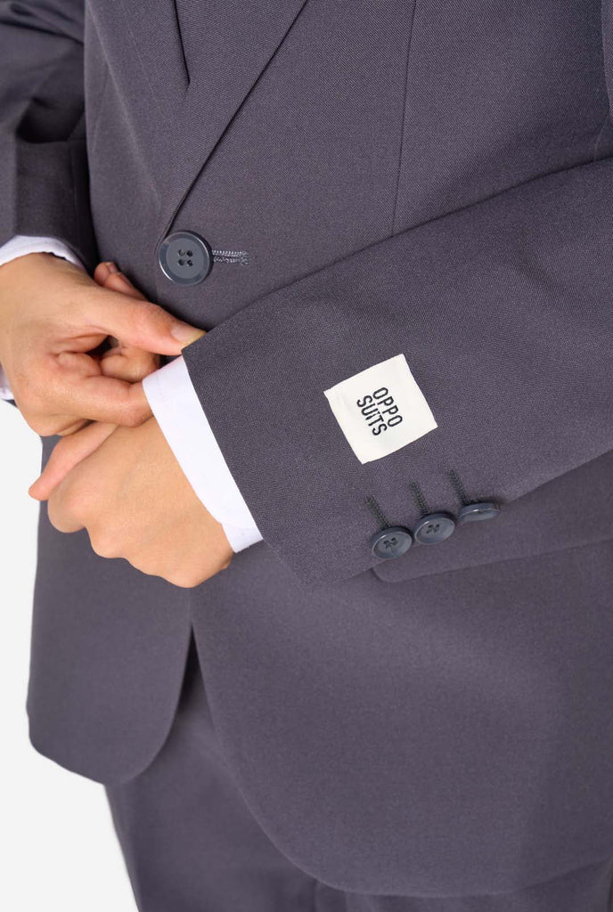 Kid wearing a stylish charcoal colored grey suit with a white dress shirt, smiling confidently. Perfect formal outfit for boys' special occasions, weddings, and parties. Sleeve close-up.
