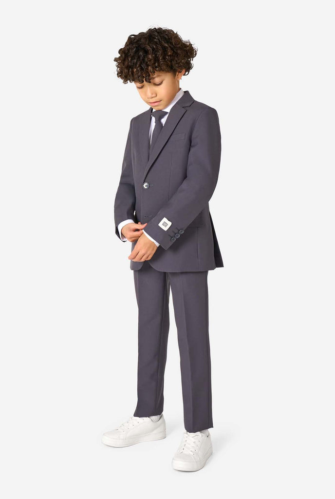 Kid wearing a stylish charcoal colored grey suit with a white dress shirt, smiling confidently. Perfect formal outfit for boys' special occasions, weddings, and parties. Full body image and looking at the floor.