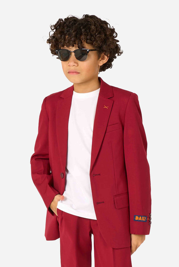 Kid wearing a stylish burgundy red suit with a white dress shirt, looking confidently and wearing sunglasses. Perfect formal outfit for boys' special occasions, weddings, and parties. Holding one hand in a pocket.