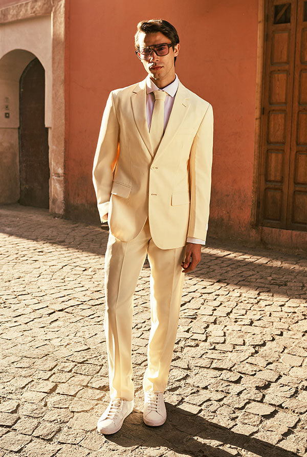 Man in a light-colored suit standing on a cobblestone street with a pink building in the background.